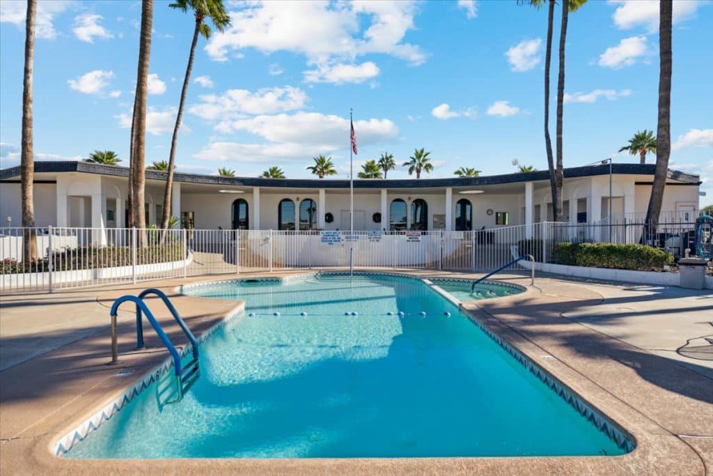 Outdoor swimming pool surrounded by palm trees and a white building with arched windows under a bright blue sky with scattered clouds in peoria az