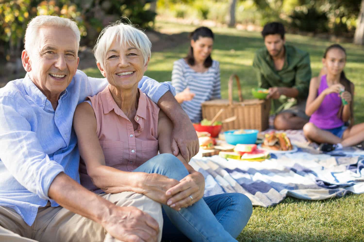 Older couple sitting on a picnic blanket, smiling at the camera, with family members blurred in the background enjoying food on a sunny day in peoria az