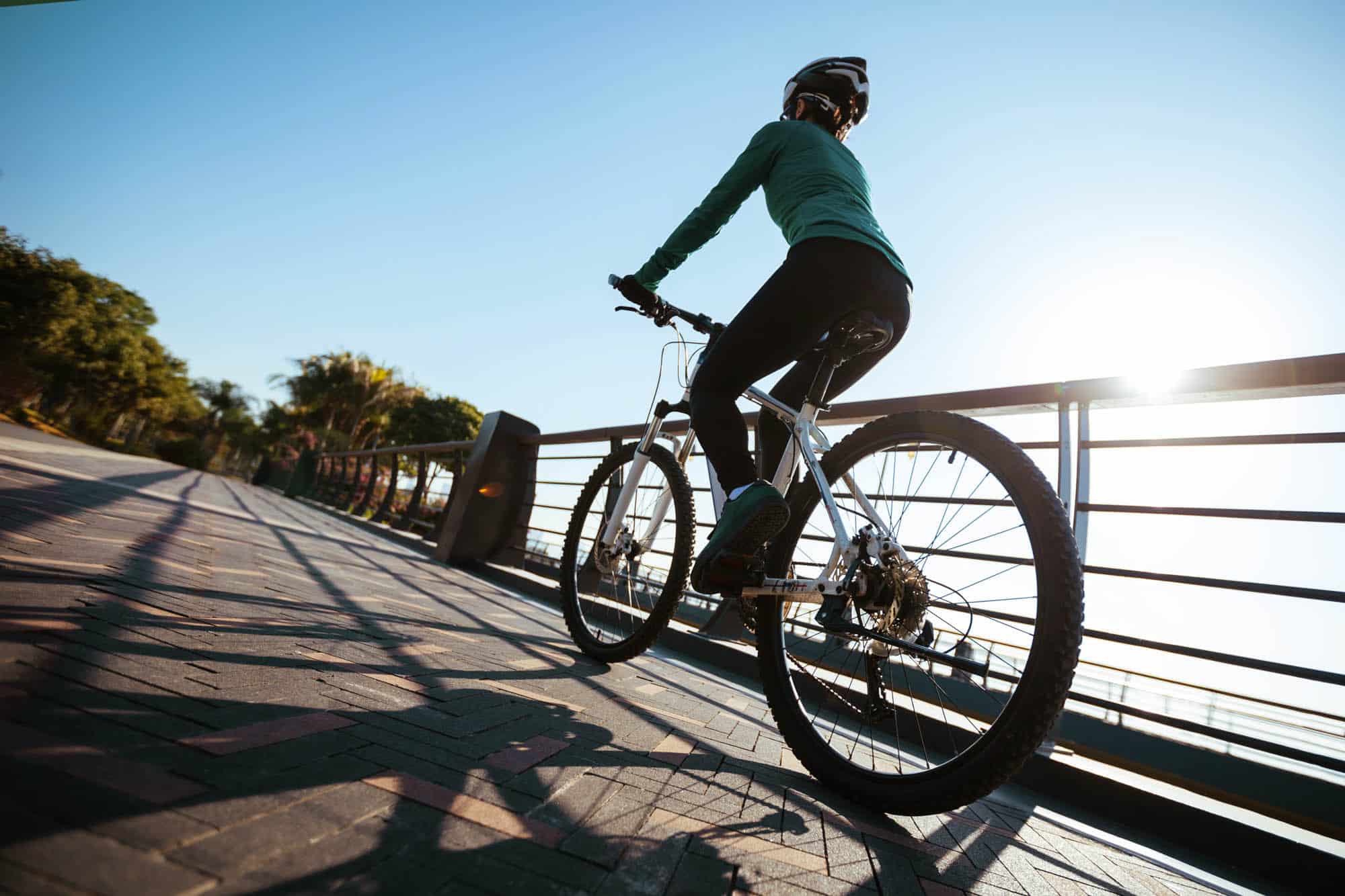 Cyclist in athletic gear riding a bike on a paved path with metal railings, trees, and sunlight shining in the background in peoria az