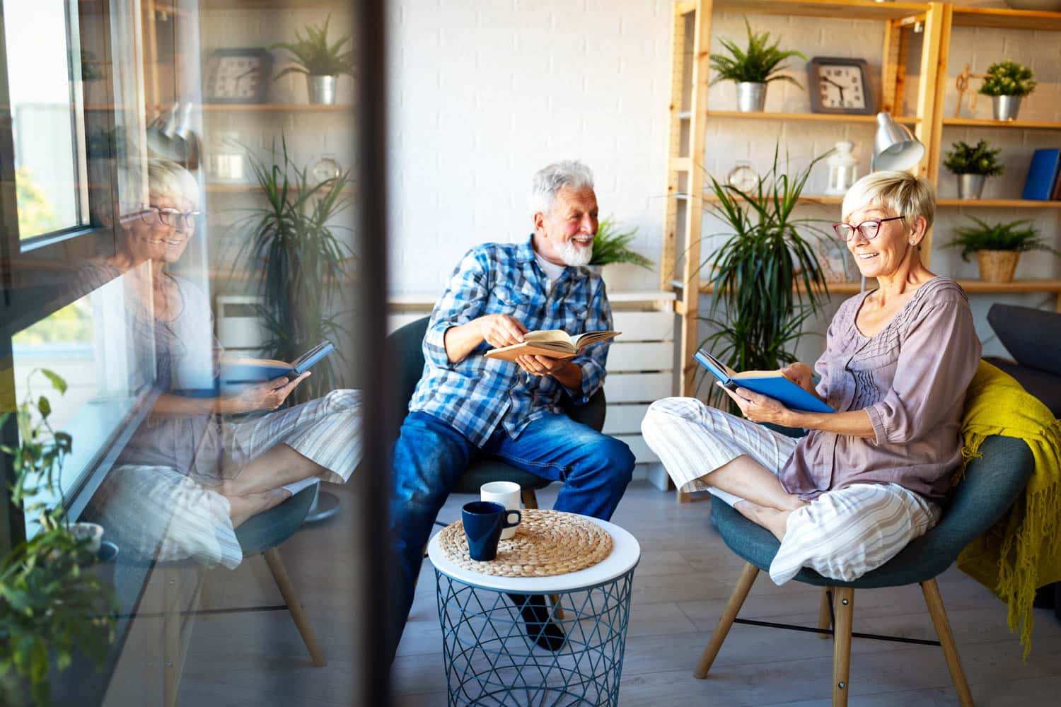 A smiling older couple sitting in a bright room, each holding a book, surrounded by plants and bookshelves in peoria az