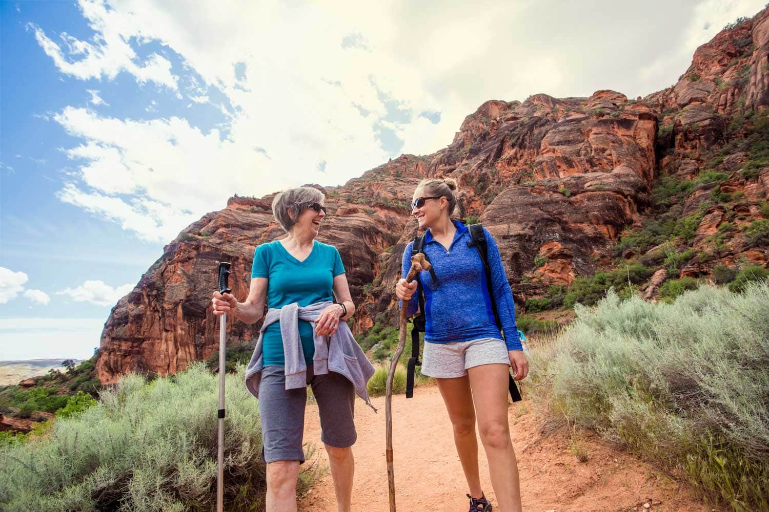 Two women, one elderly and one younger, hiking on a trail with rugged red rock formations in the background, smiling and carrying walking sticks in peoria az